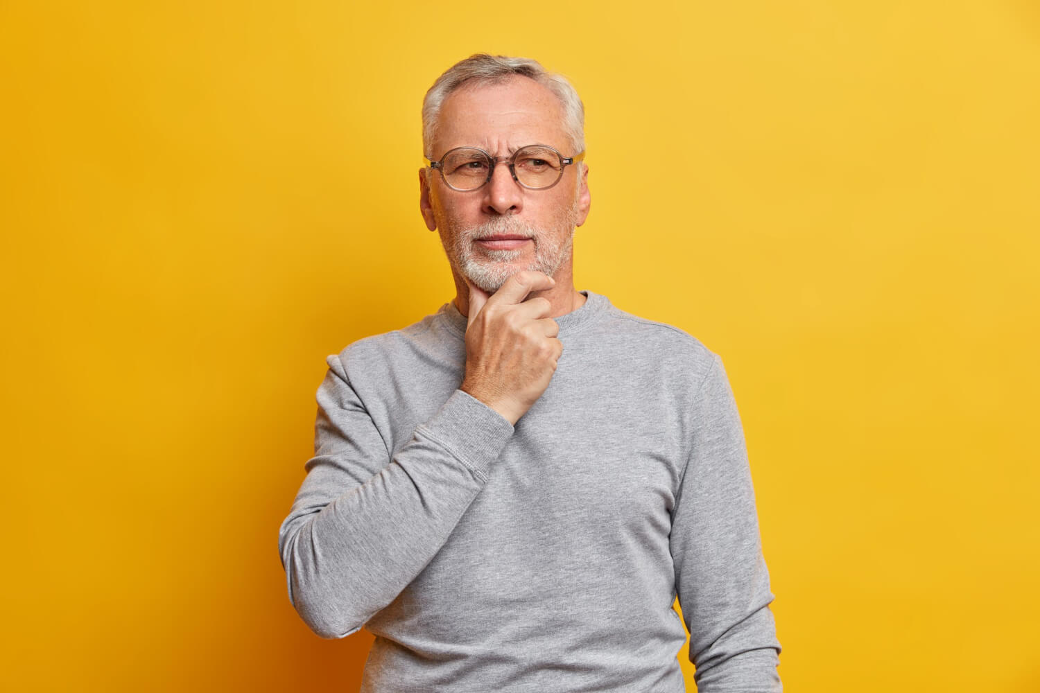 Homem idoso de óculos e barba grisalha com expressão pensativa sobre implante dentário em Santo André, contra fundo amarelo vivo.