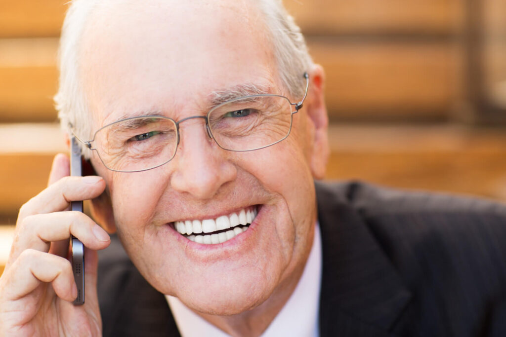 Homem idoso de terno e óculos sorri confiante enquanto fala ao celular, exibindo dentes saudáveis. Exemplo de reabilitação oral na Clínica Adachi em Santo André.