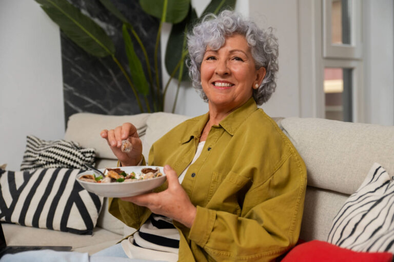 Mulher idosa sorridente segurando prato de comida, demonstrando segurança ao mastigar com prótese protocolo na Clínica Adachi em Santo André.