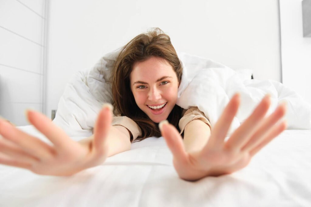 Mulher sorrindo deitada na cama e esticando os braços. Saiba se quem tem bruxismo pode usar facetas de porcelana em Santo André.
