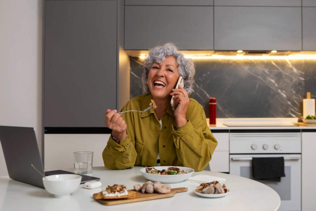 Mulher sênior sorridente comendo na cozinha enquanto fala ao celular, demonstrando a segurança da prótese protocolo em Santo André.